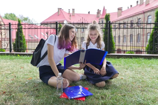 Two schoolgirls sitting on grass with books near school building, girls sisters, teenager and elementary school student laugh and look at textbooks