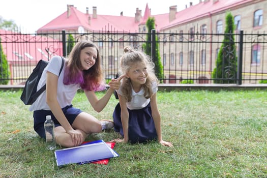 Two schoolgirls sitting on grass with books near school building, girls sisters, teenager and elementary school student laughing