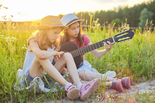 Children sitting in nature with classical guitar, two girls learning playing the guitar and singing, enjoying music and summer day