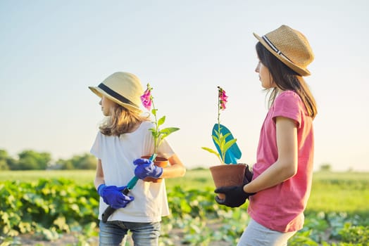 Portrait of two girls children with flowers in pots, gloves, with garden shovels. Young gardeners in hats, background rural nature, landscape, sky, spring summer season