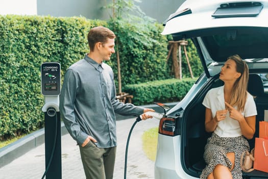 Young couple travel with EV electric car charging in green sustainable city outdoor garden in summer shows urban sustainability lifestyle by green clean rechargeable energy of electric vehicle innards