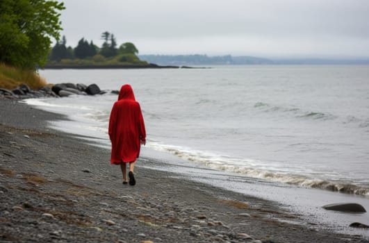 A lone figure in a vibrant red cloak walks down a pebble-covered shoreline, with the calm sea to one side and a line of trees to the other, under an overcast sky