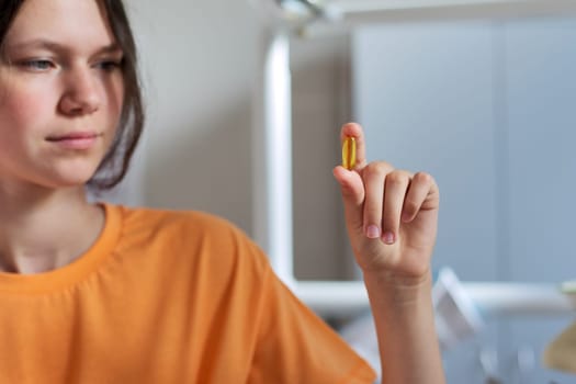 Teenage girl holding yellow vitamin d gel capsule in hand, dentist office background. Nutritional supplements, vitamins, healthy nutrition of adolescents and young people