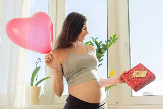 Happy young beautiful pregnant woman with gift box and red heart balloon, gift for Valentines day, for spring holidays. Motherhood and pregnancy
