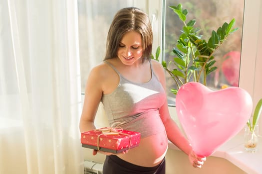 Happy young beautiful pregnant woman with gift box and red heart balloon, gift for Valentines day, for spring holidays. Motherhood and pregnancy