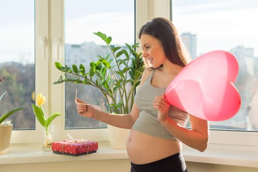 Young beautiful pregnant woman with gift box and red balloon heart, female at home near window, Valentines day and holidays. Motherhood and pregnancy