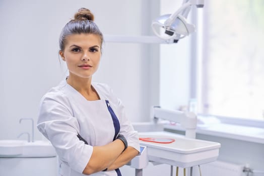 Young confident female dentist doctor with folded arms, dentistry cabinet background, copy space