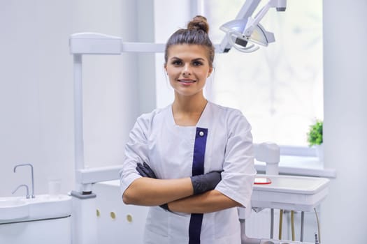 Portrait of young confident smiling dentist doctor woman, female with arms crossed looking at camera standing in dentistry office