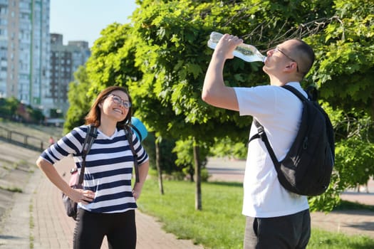 Mature smiling man and woman in sportswear with backpacks exercise mat walking in city park talking drinking water from bottle, active healthy lifestyle of middle-aged people