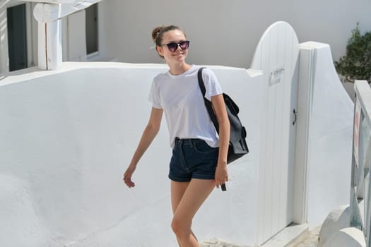 Walking smiling young beautiful girl in shorts, sunglasses, backpack, female tourist in sunny summer city with white Mediterranean architecture, copy space