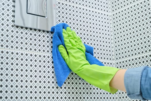 Woman in gloves with rag doing cleaning in bathroom, cleaning and polishing chrome toilet button on tiled wall