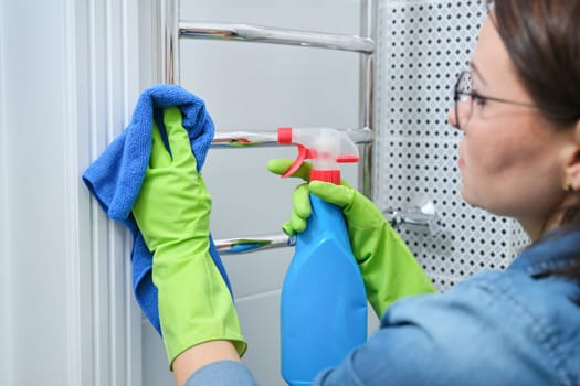 Cleaning bathroom, woman in gloves with rag detergent cleaning polishing chrome heated towel rail