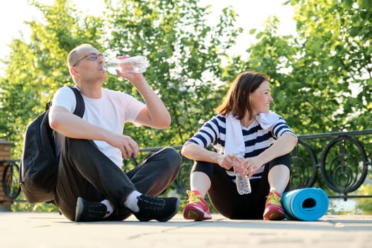 Relationship between middle-aged people, couple man and woman 40 years old talking, relaxing after fitness in park drinking water from bottle, active lifestyle