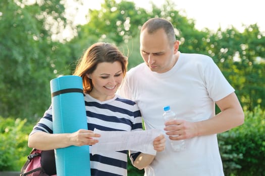Mature couple reading paper document letter. Man and woman outdoor in sportswear with exercise mat. Healthy active lifestyle of middle-aged people.
