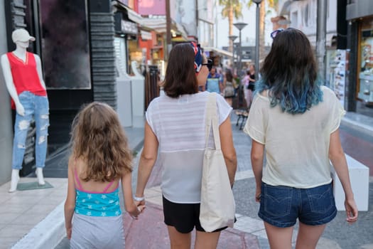 People mother and two daughters walking along street of small resort town, family vacations together, summer fun shopping in souvenir shops, back view