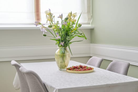 Home dining room interior, table with white tablecloth, spring summer bouquet of flowers in vase, tray with ripe strawberries