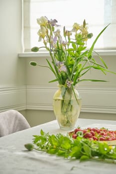 Home dining room interior, table with white tablecloth, spring summer bouquet of flowers in vase, tray with ripe strawberries