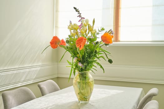 Bouquet of spring summer flowers and red poppies in vase, standing on table in living room, dining area near window, copy space