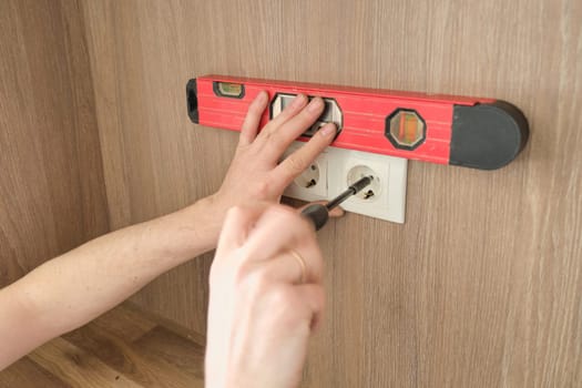 Repair and installation of furniture, hands of electrician worker installing electrical outlet in furniture using screwdriver and level