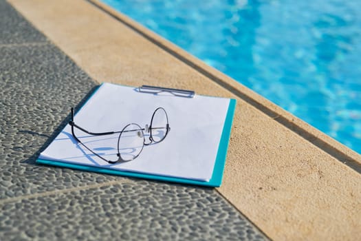 Sports concept, active healthy lifestyle, business hotel. Nobody, glasses, blank paper, clipboard near outdoor swimming resort pool