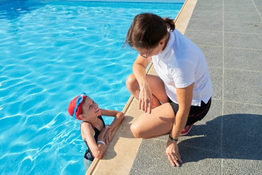Active healthy lifestyle in children. Woman trainer talking to girl child in sports swimsuit hat and goggles for swimming near outdoor pool