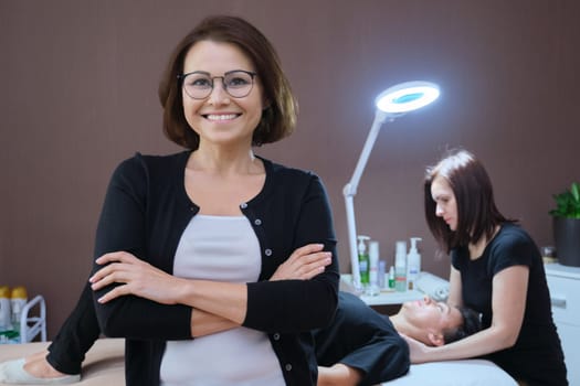 Portrait of beauty salon owner woman, posing mature confident smiling female, background massage procedure on the table