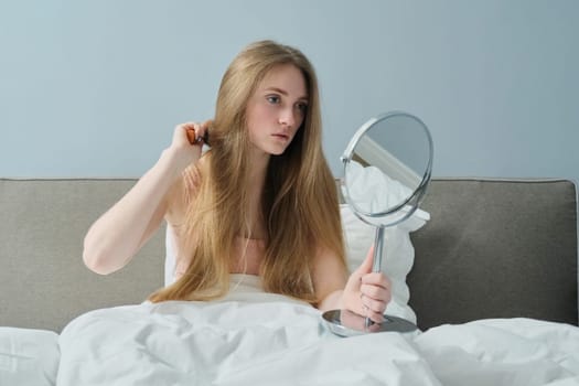 Young beautiful girl combing long hair with brush and mirror. Female sitting in bed, hair beauty care health