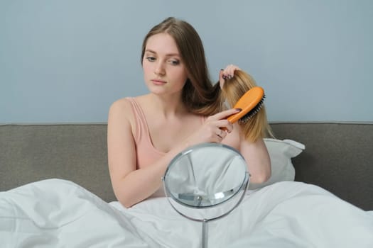 Young beautiful girl combing long hair with brush and mirror. Female sitting in bed, hair beauty care health