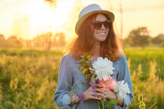 Happy beautiful young teen girl on meadow in glasses hat dress with bouquet of flowers. Summer holidays, nature, beauty. Background sky, field, golden hour, copy space