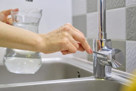 Woman pouring water into jug from kitchen faucet, closeup of hand on chrome mixer