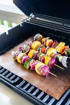 Skewered beef and fresh veggies sizzle on a copper grill mat over a gas outdoor grill.