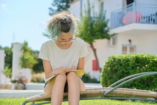 Student girl 15, 16 years old sitting outdoor, writing in school notebook, female studying at home