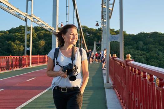 Positive middle-aged woman walking on bridge. Mature female with backpack photo camera smiling on sunny summer day