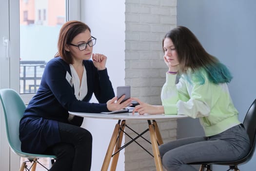 Female psychologist talking with teenage girl 15, 16 years old. Teenager telling and showing on smartphone problems. Mental health of youth, adolescence