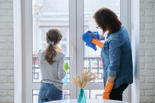 Mother and daughter child in rubber gloves with detergent and rag washing windows together. Girl helping woman do house cleaning