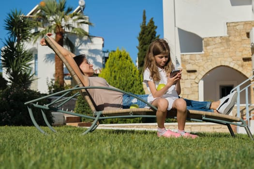 Resting happy family mother and daughter sitting in an outdoor chair on sunny summer day. Mother relaxing with closed eyes, girl playing on smartphone