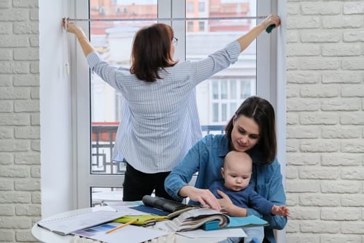 Window decoration with curtains, woman designer taking window sizes. At the table, sitting mother and baby choosing fabrics, color, model