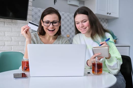 Family, mother and teenage daughter using laptop computer credit card and doing online shopping. Women sitting at home in kitchen talking smiling choosing gifts
