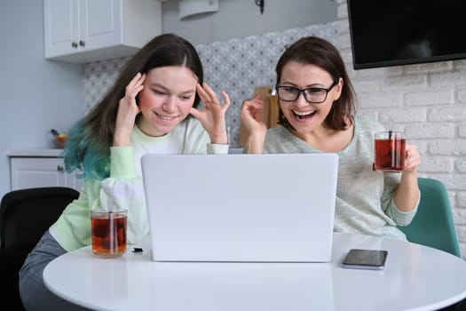 Relations between mother and teen daughter, parent and teenager sitting at home in kitchen drinking tea together and looking at laptop monitor, watching videos and laughing
