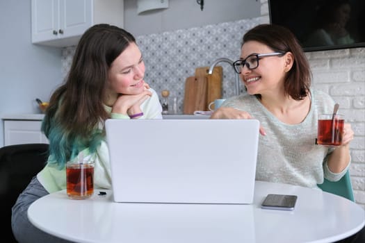 Communication mother and daughter teenager, talking parent and child sitting at home in kitchen and looking at laptop screen
