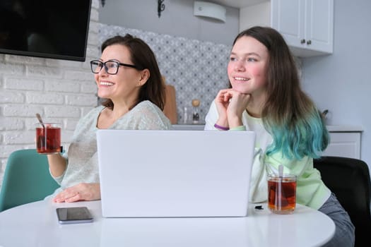 Communication mother and daughter girl teenager, at home in kitchen sitting at table with laptop computer, drinking tea, smiling looking out the window