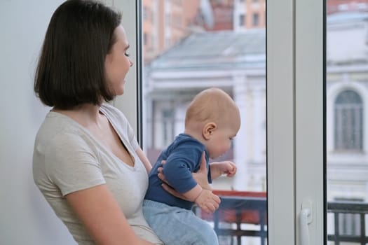 Portrait of young beautiful mother and toddler son, woman holding baby in her arms standing at home near the window, copy space