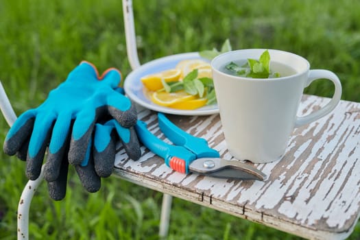 Close-up of white vintage chair on green grass in garden. With garden secateurs pruner, gloves, cup of fresh herbal mint tea with lemon