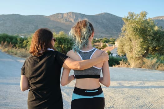 Athletic healthy active family, mother and teenage daughter walking in sportswear on the road, view from the back. Mountains background, summer nature