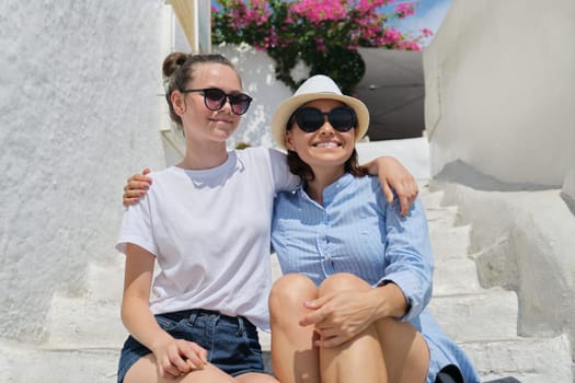 Happy tourists mother and teen daughter together hugging sitting on white steps enjoying scenery, summer family vacation, white Mediterranean architecture