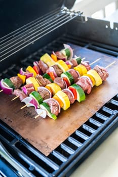 Skewered beef and fresh veggies sizzle on a copper grill mat over a gas outdoor grill.