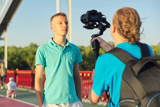 Young man model and male operator taking photos videos with camera and stabilizer, filming backstage, background foot bridge over the river on sunny summer day