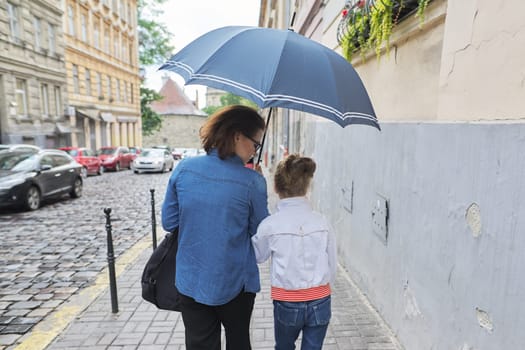 Rainy autumn weather in city, woman with child girl walking under an umbrella in street, rear view