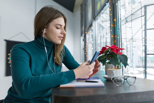 Young woman listening in headphones using smartphone for work and study, autumn winter season in cafe with cup of hot drink