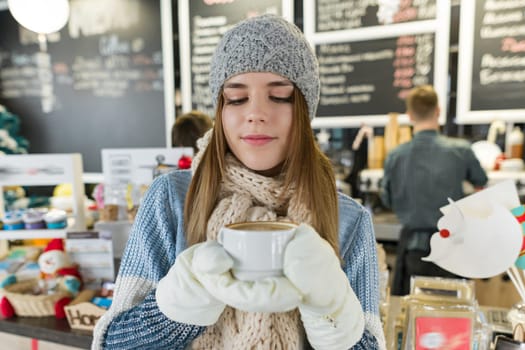 Winter portrait of young beautiful woman in knitted scarf, knitted hat, mittens, warm sweater with cup of coffee. Girl near bar counter in coffee shop